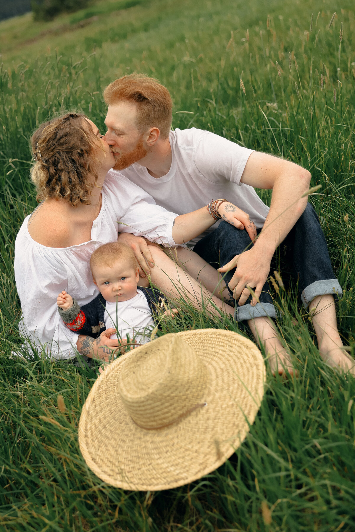 Summer Family Photo of Parents Kissing with Baby Sitting in Meadow and Straw Hat in Foreground