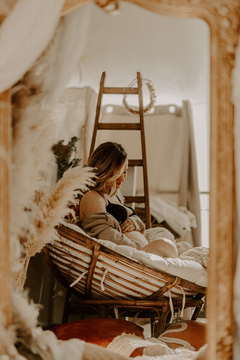 Femme enceinte, assise dans une chaise ronde en bois garnie d'un gros coussin posant dans un décor cosy pour une séance photo grossesse.