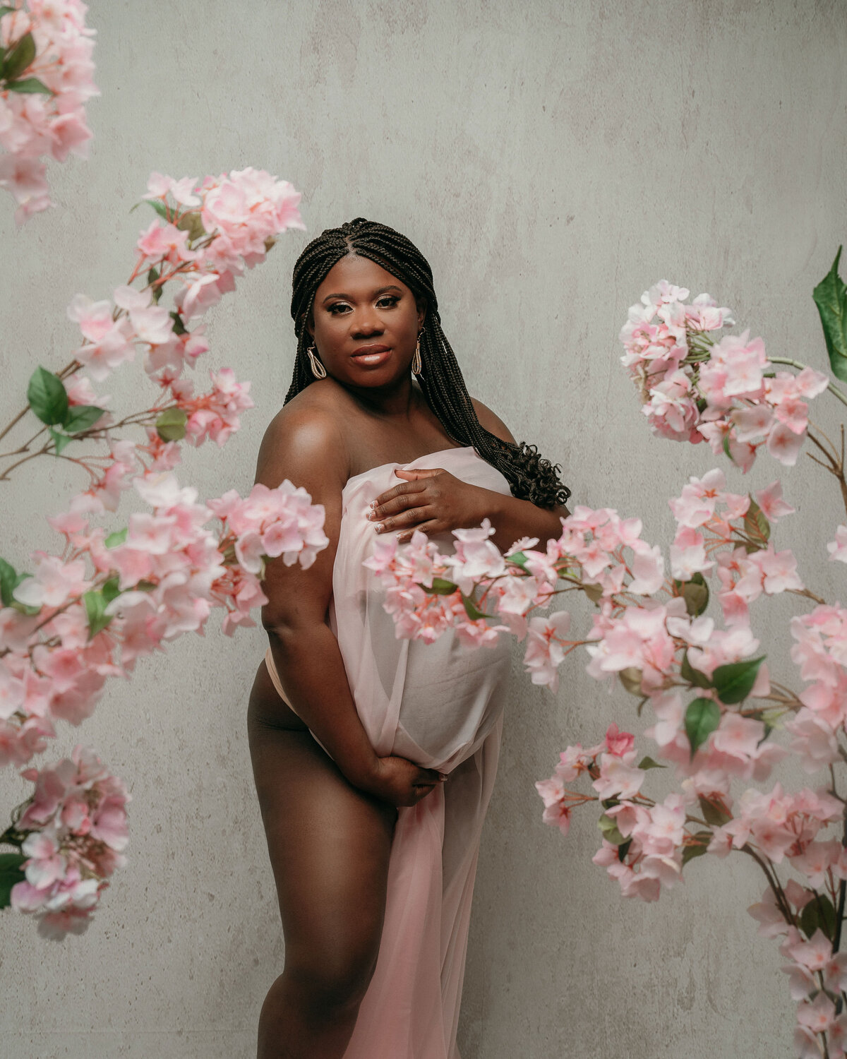 Pregnant woman nude wrapped in silk fabric holding her baby bump on off white backdrop and pink flowers all around her