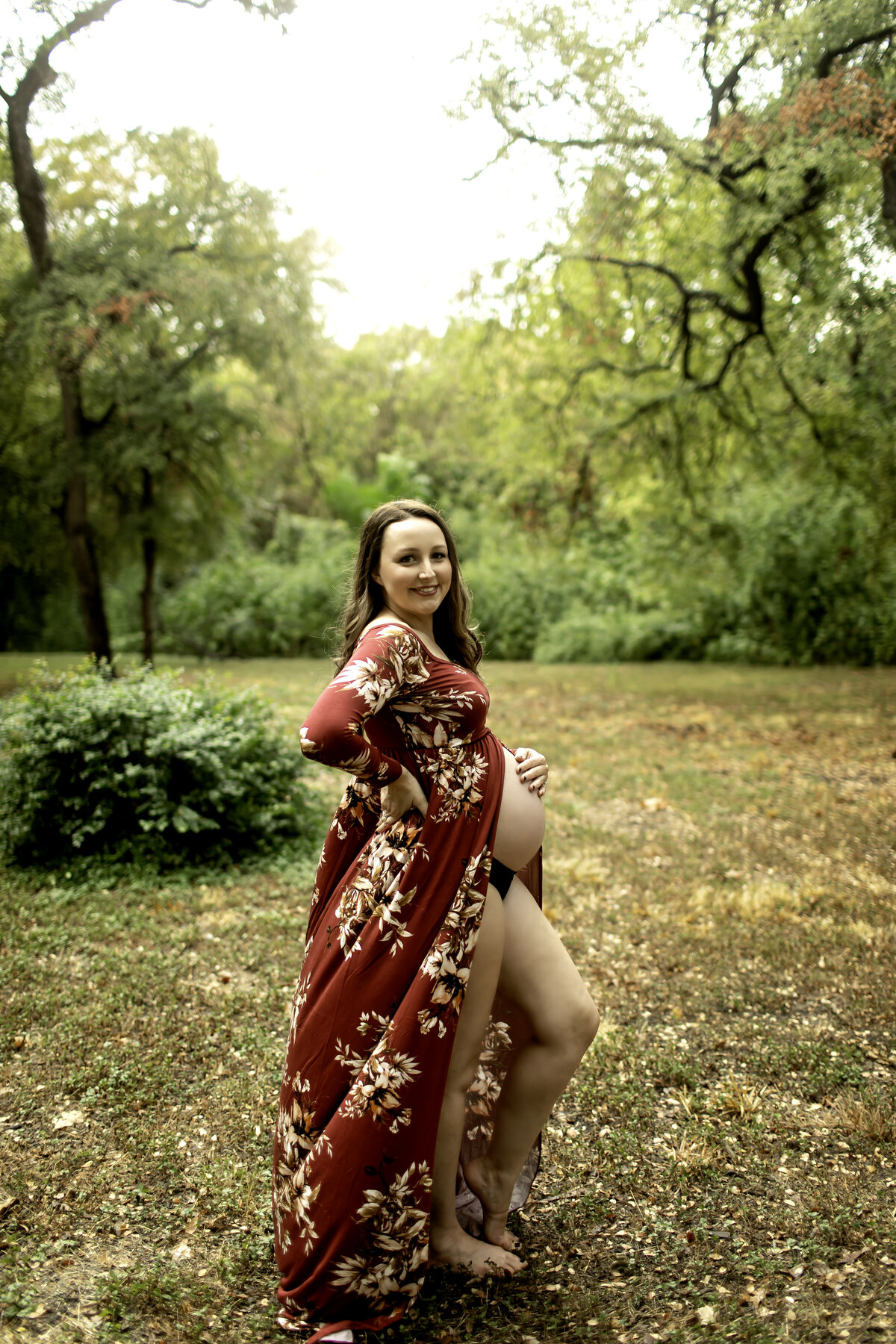 A striking maternity photo of the expectant mom in a red dress, standing gracefully at Trinity Park, with the lush green trees and serene backdrop, captured by Chunky Monkey Photography.