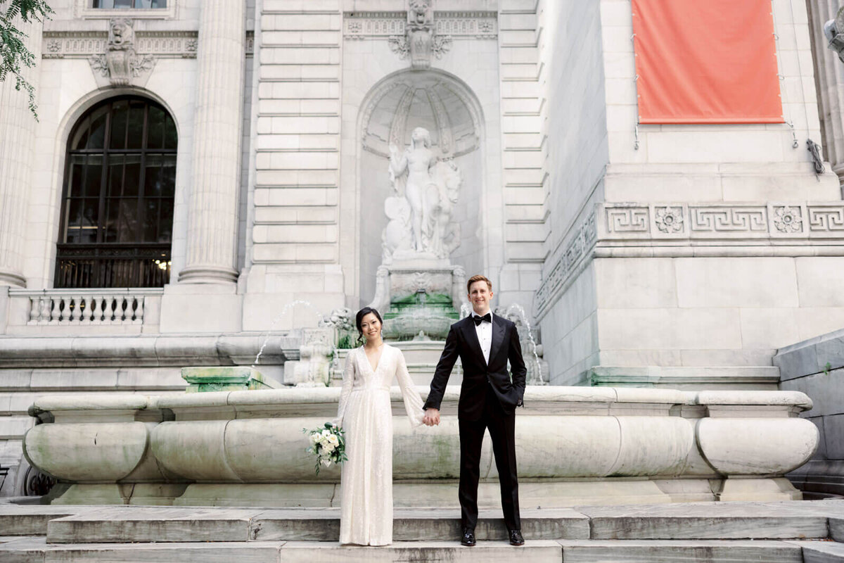 The bride and the groom are smiling in front of a fountain outside New York Public Library Park. Image by Jenny Fu Studio
