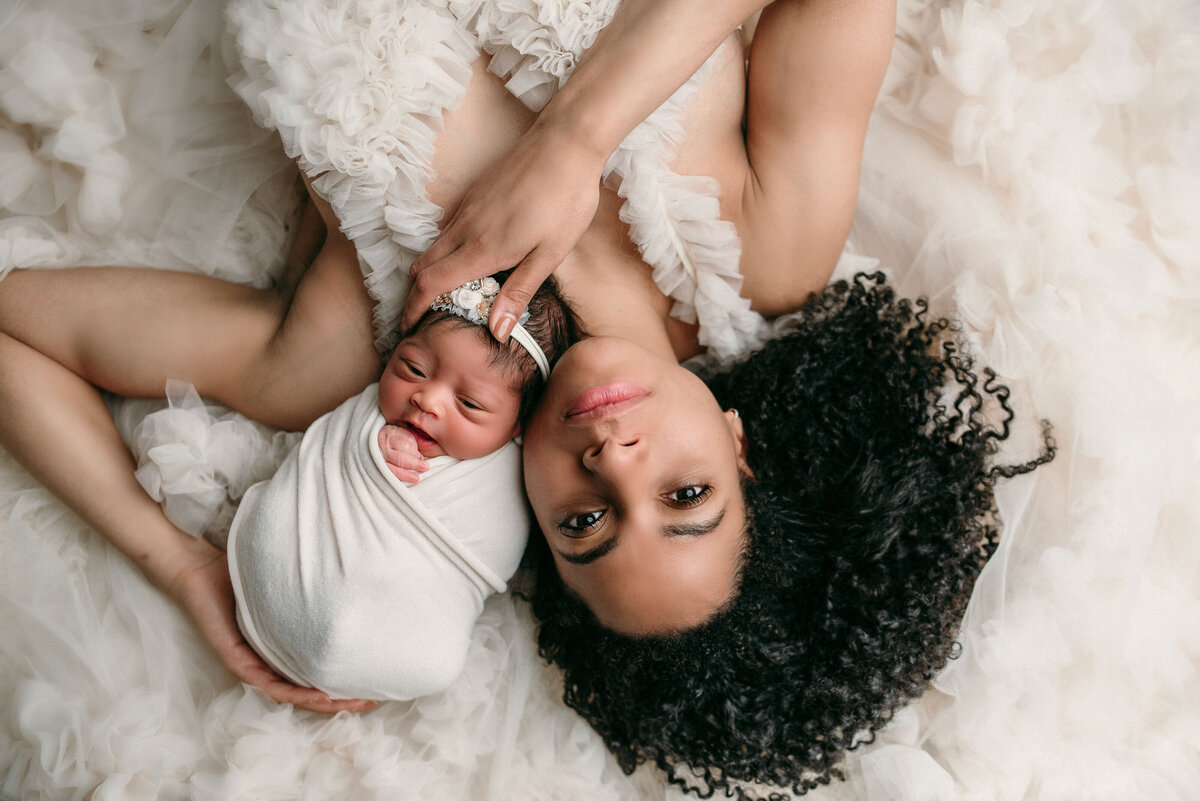 Long brown haired woman in white gown laying on back holding her newborn baby girl swaddled in white fabric