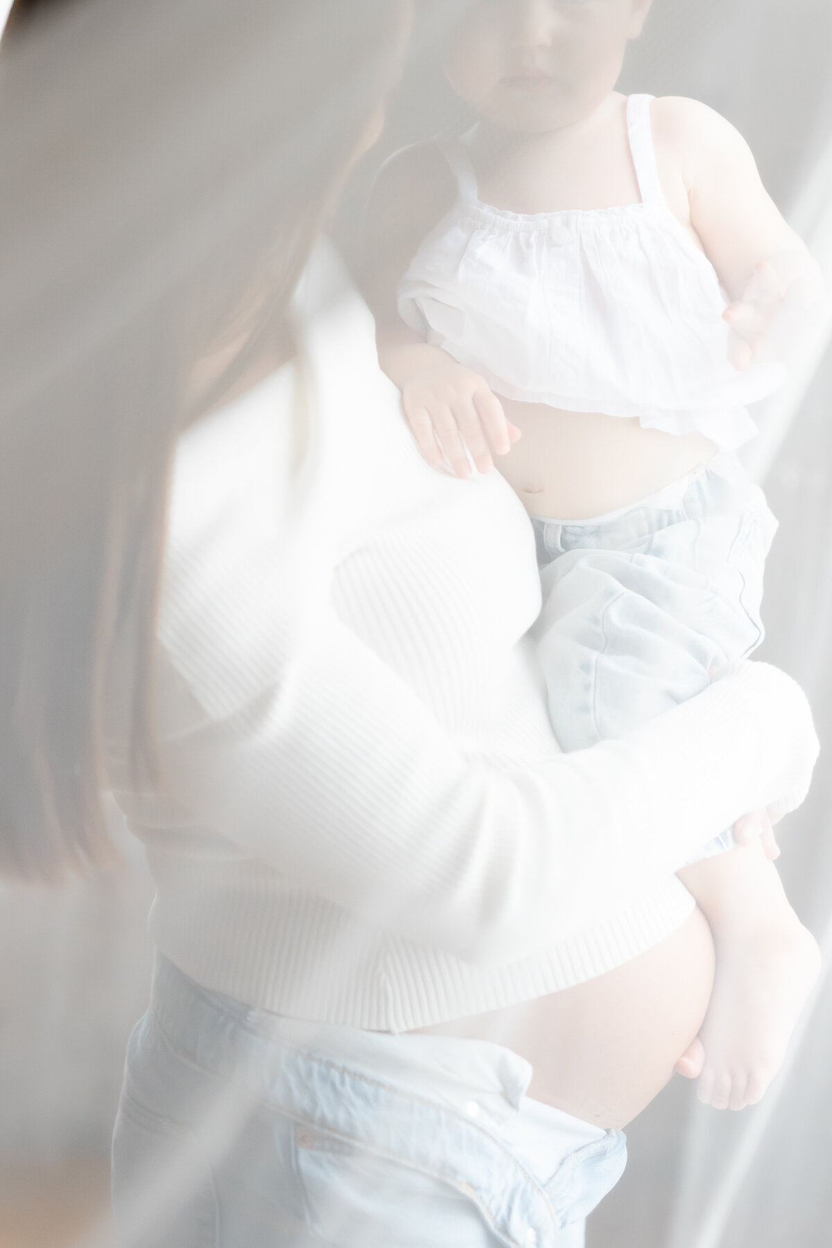 a pregnant mother holds onto her toddler over her pregnant belly as a sheer curtain sweeps in front of them.