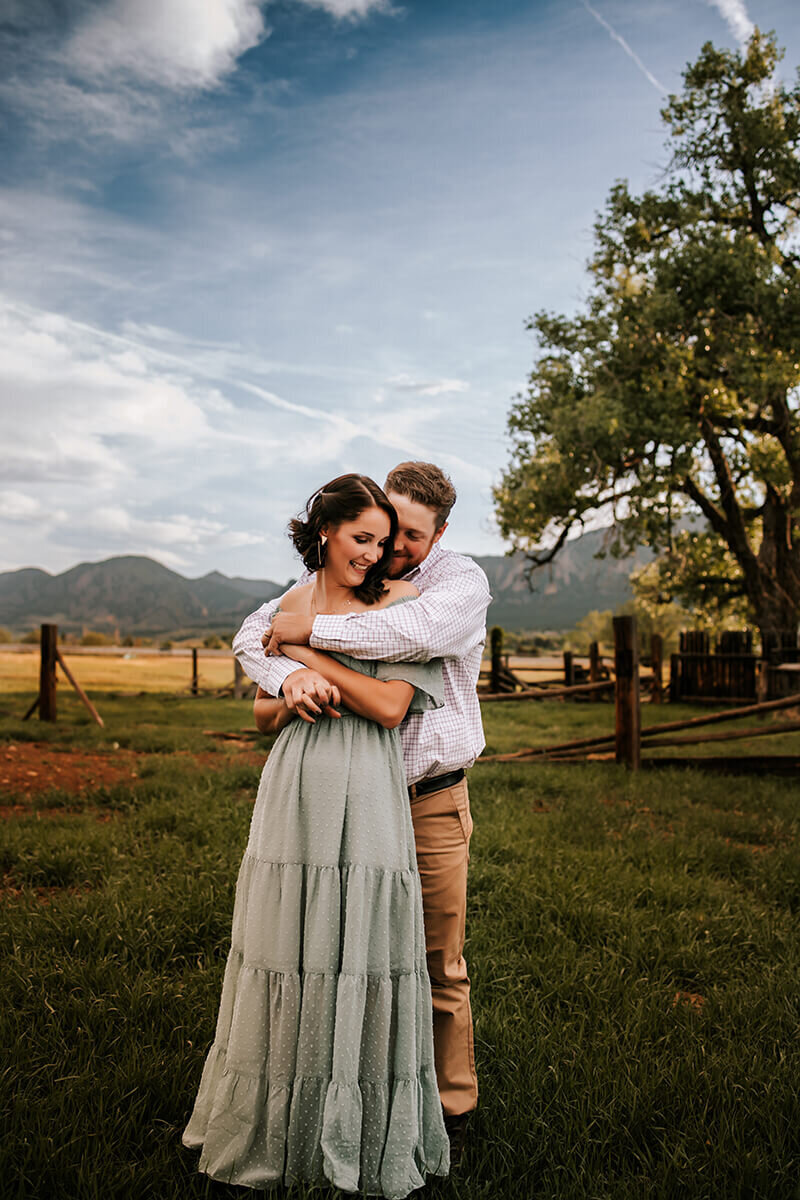 Husband hugs wife from behind while wearing a shoulder less green dress in a grassy field in Boulder, Colorado