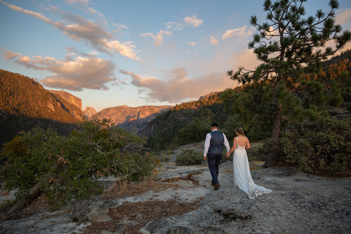 A bride and groom walk hand in hand along a granite slab in Yosemite with Half Dome and El Capitan glowing orange in front of them.