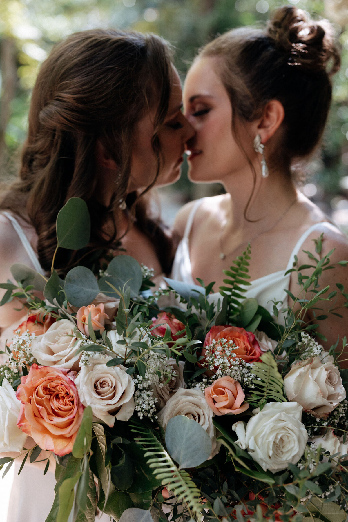 Two brides kiss as they hide behind their peach, pink, and greenery bouquets.