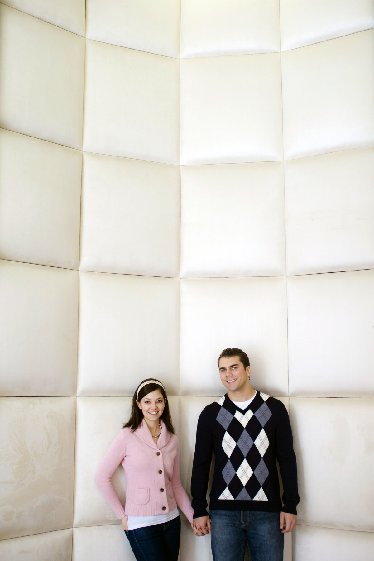 A couple standing in front of a cream padded wall smiling 