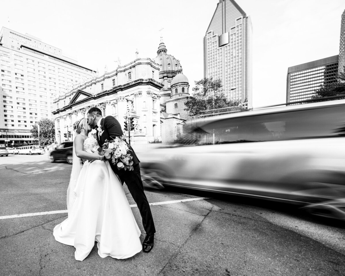 A timeless black-and-white photo capturing a couple sharing a passionate kiss on the sidewalk, with blurred movement of the busy street behind them, highlighting their moment of love amidst the hustle and bustle of the city.