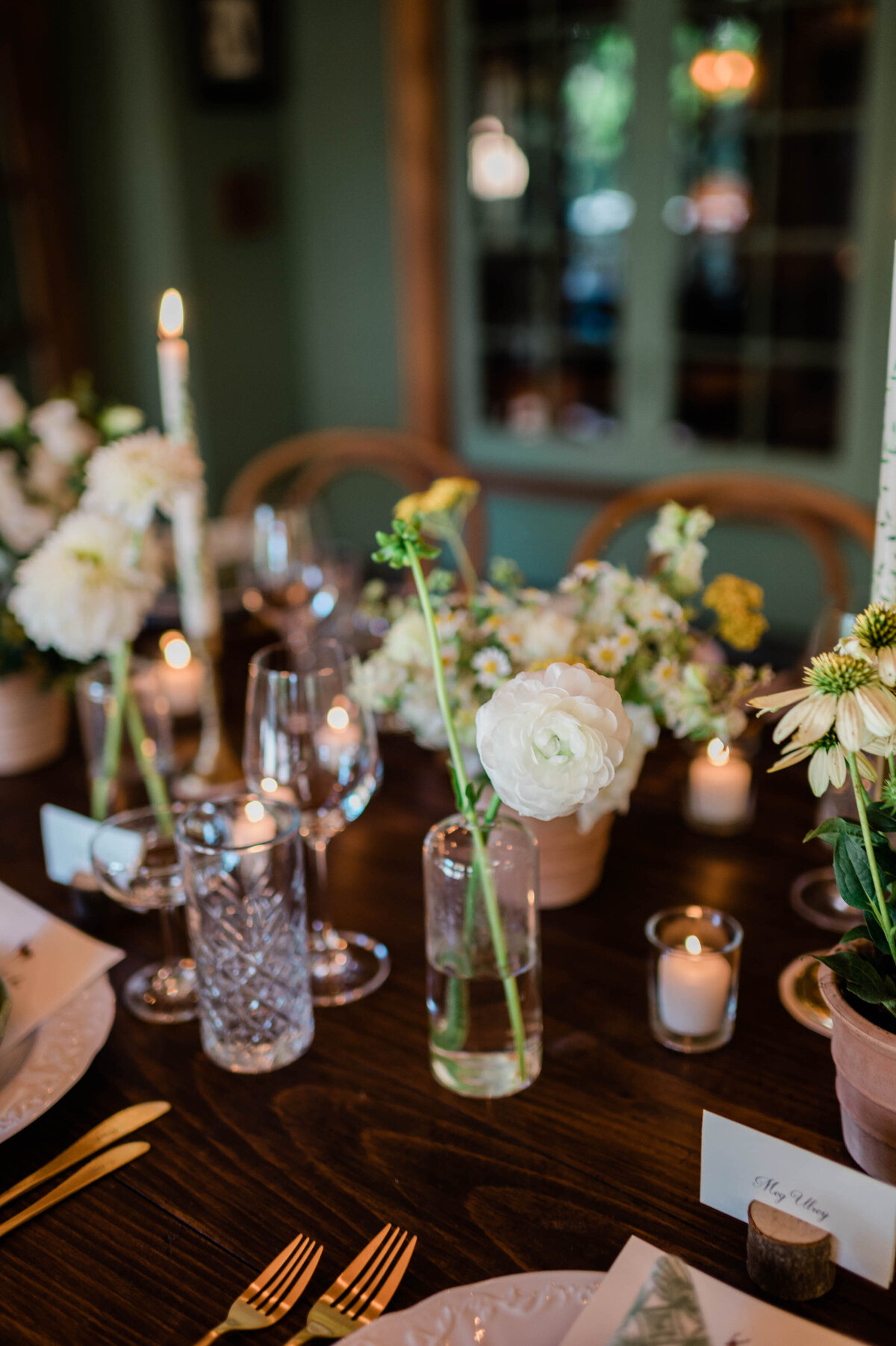 A delicate white flower in a glass vase sits on a wooden table, surrounded by candles, wine glasses, and other floral arrangements.