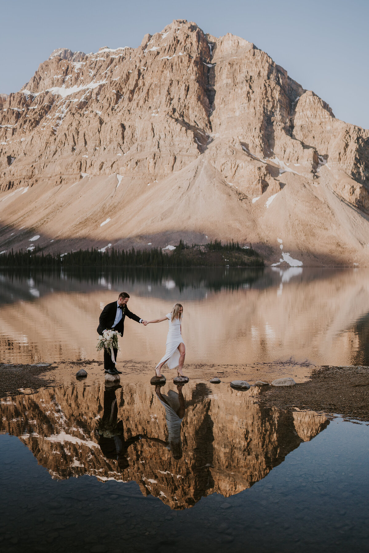 Bride leads groom over rocks at Bow Lake during sunrise as the lake reflects