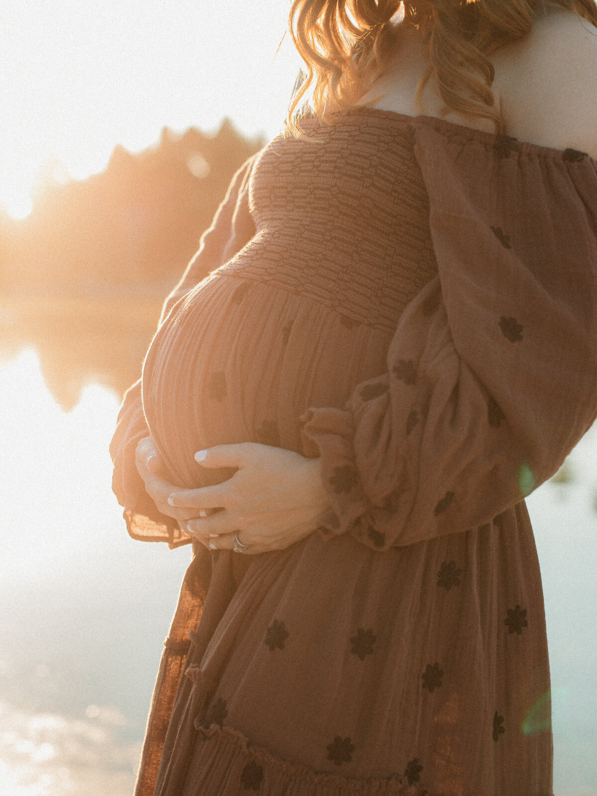Close-up of a pregnant belly in a rust-colored off-shoulder dress, softly backlit by sunset near a lake.
