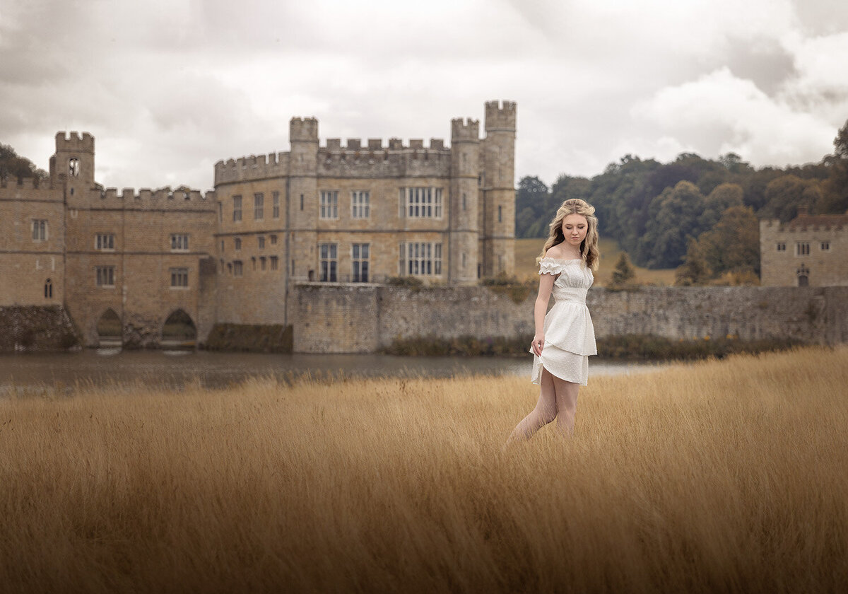 Image of a high school senior wearing a white dress and walking through the long grass fields in front of Leeds Castle, Kent. Taken by Norwich Senior portrait photographer Claire Howes.