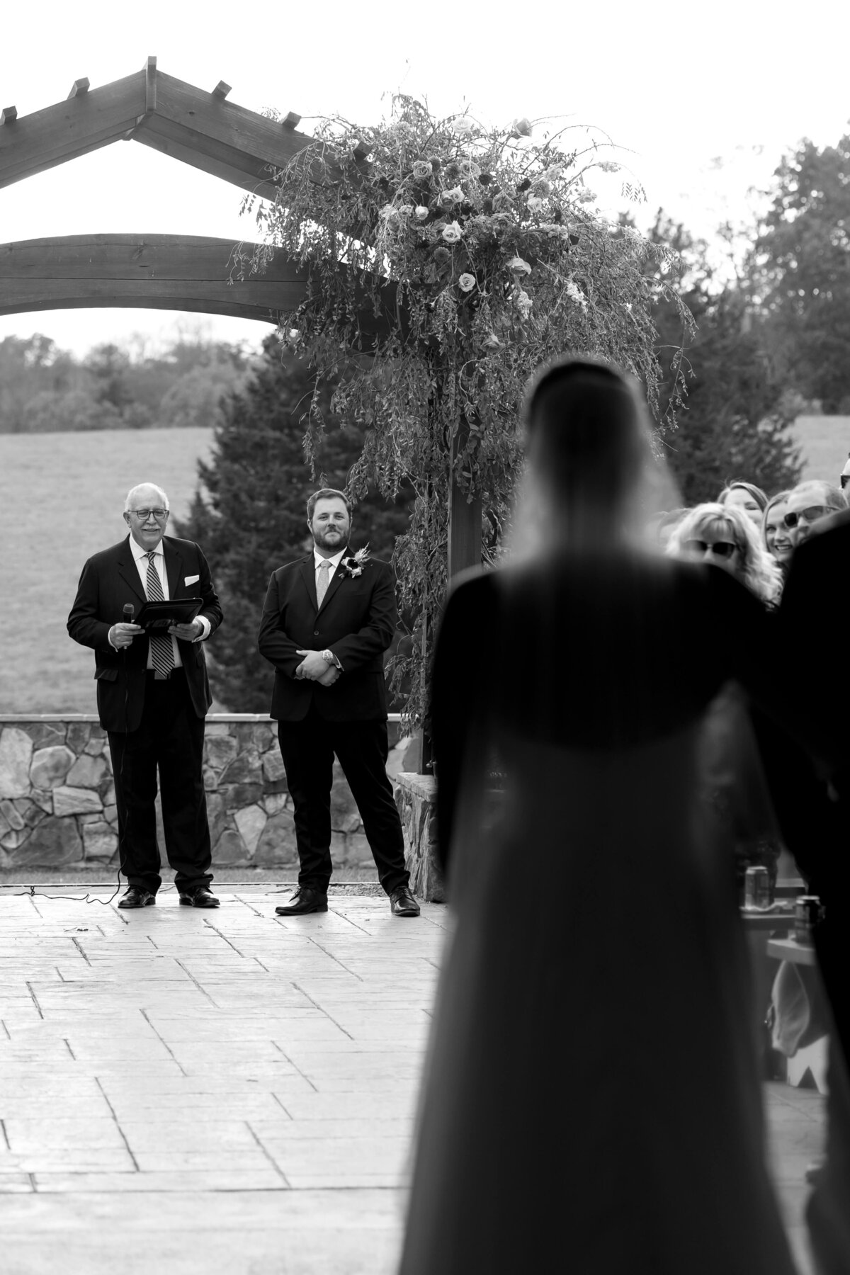 black and white wedding picture with groom standing with the officiant as his bride enters the ceremony space with her father for their Shenandoah National Park wedding