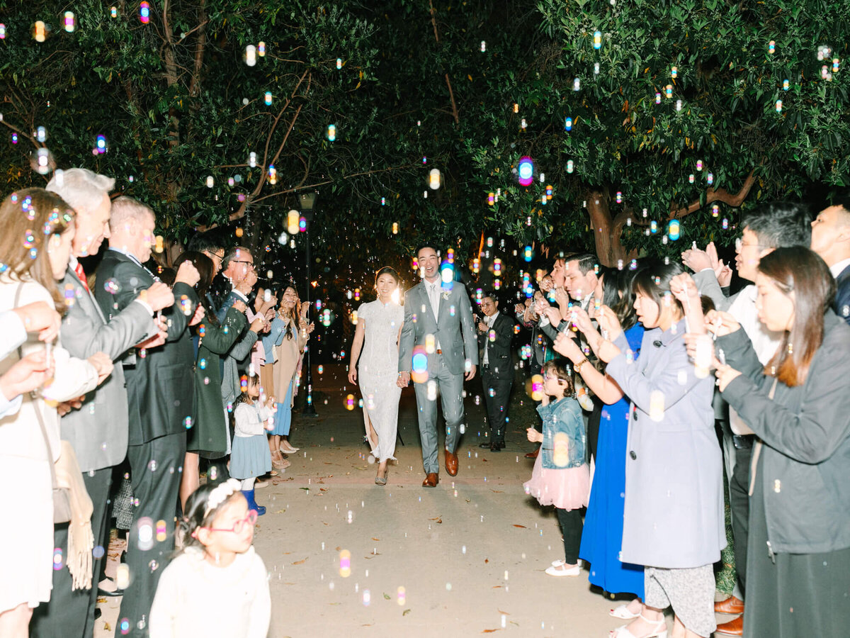 A couple in formal attire walks joyfully through a path lined with people blowing bubbles, under a canopy of trees at a nighttime celebration.
