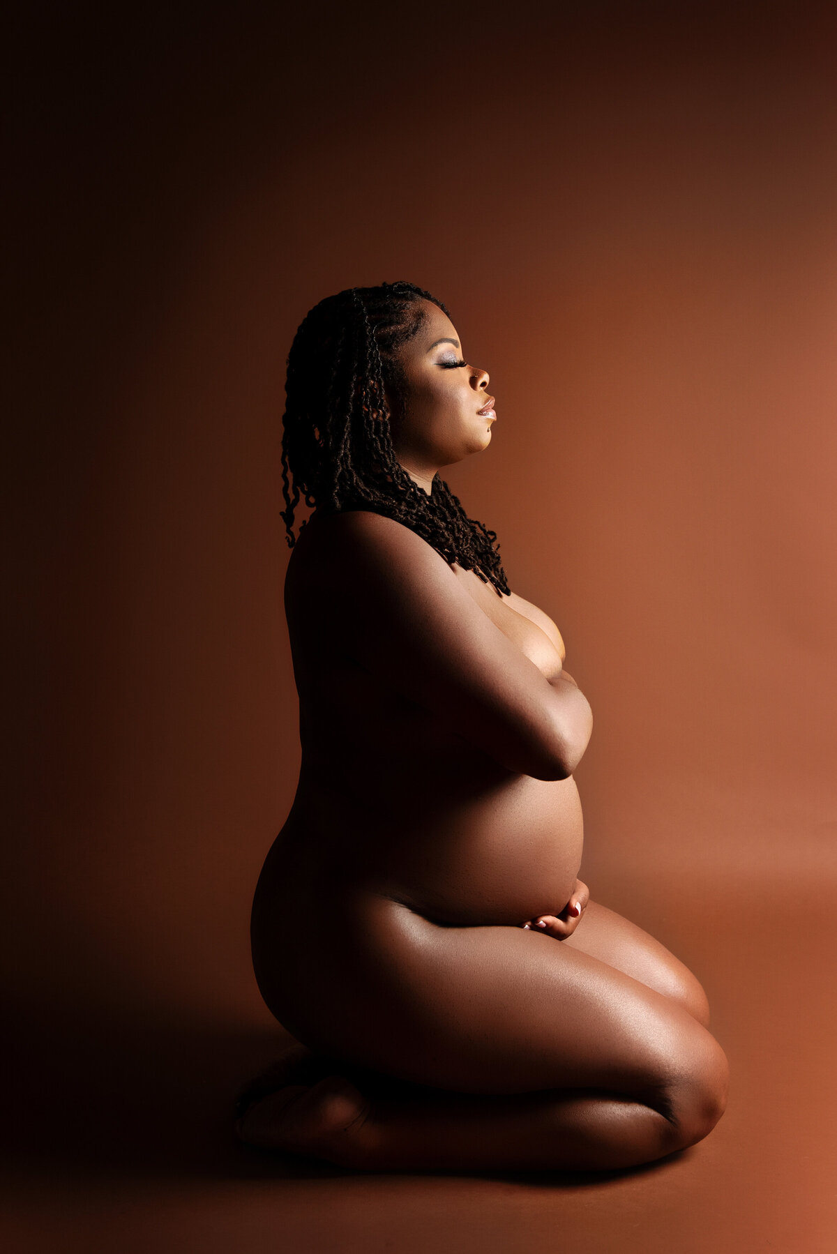 st-louis-maternity-photographer-nude-pregnant-mom-kneeling-facing-to-the-side-with-brown-background-and-dramatic-light