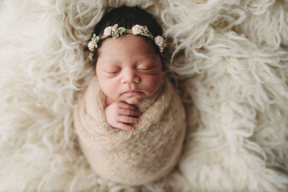 Baby newborn girl in cream knitted swaddle and white flower headband laying on cream fur mat