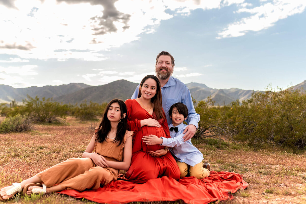 Family sitting in the Arizona desert, surrounding expecting mom with love and hugs.