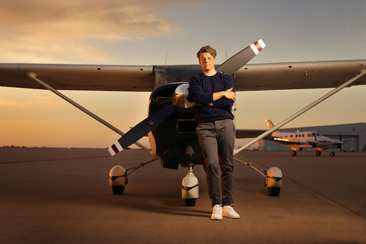 A senior stands confidently next to a small aircraft, showcasing their passion for aviation at sunset. Captured by a Dallas senior photographer.