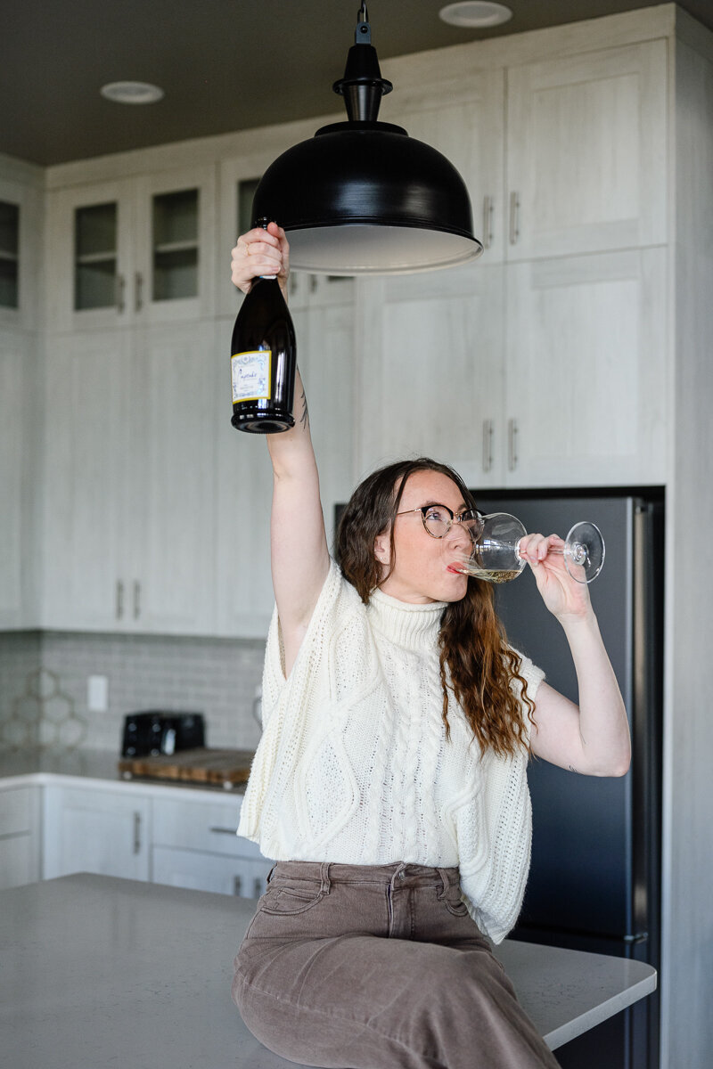 A brand photographer in Denver takes a photo of a woman sitting on a kitchen counter holding up a bottle of wine and celebrating during a commercial photoshoot in Denver for branding photos.