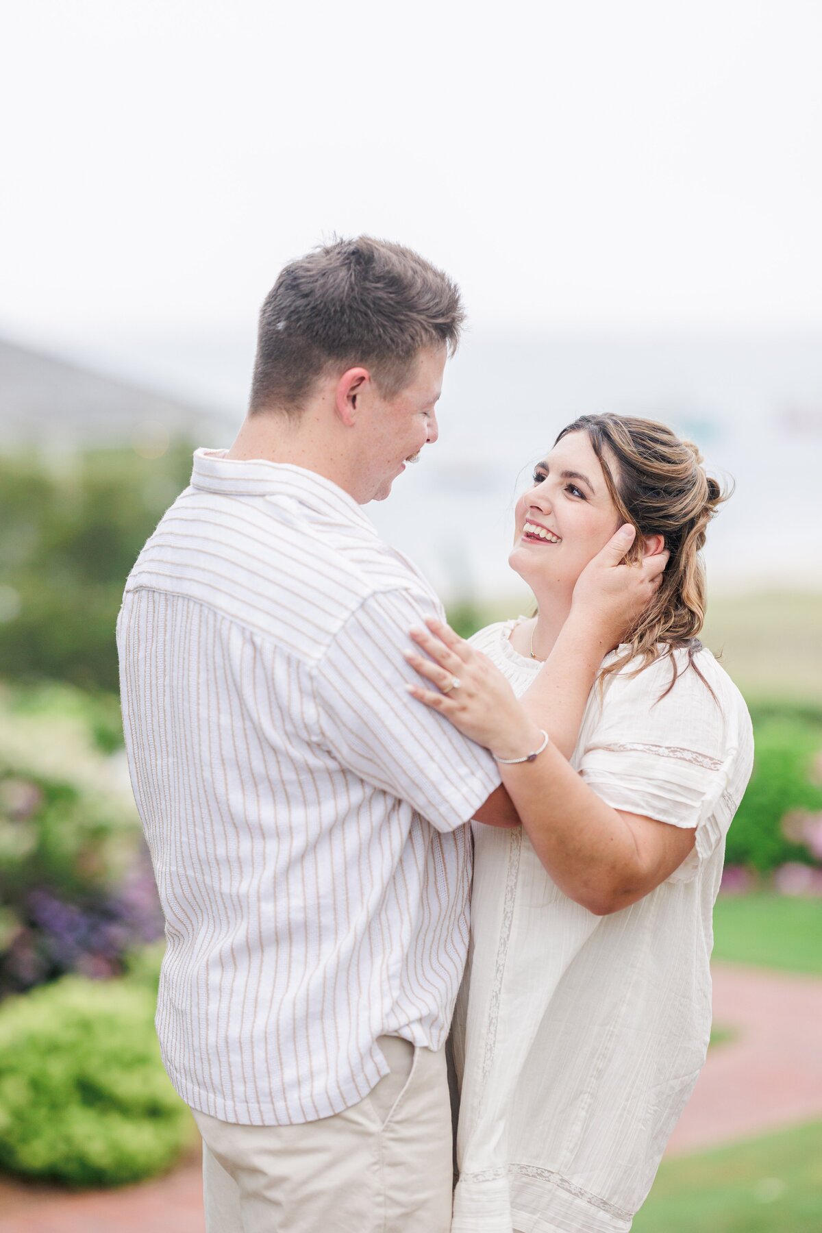 Close-up of a couple gazing at each other and smiling, the man’s hands gently touching the woman’s face, with blurred greenery in the background at Chatham Bars Inn in Chatham, Massachusetts — representing the portfolio of a Cape Cod engagement photographer.