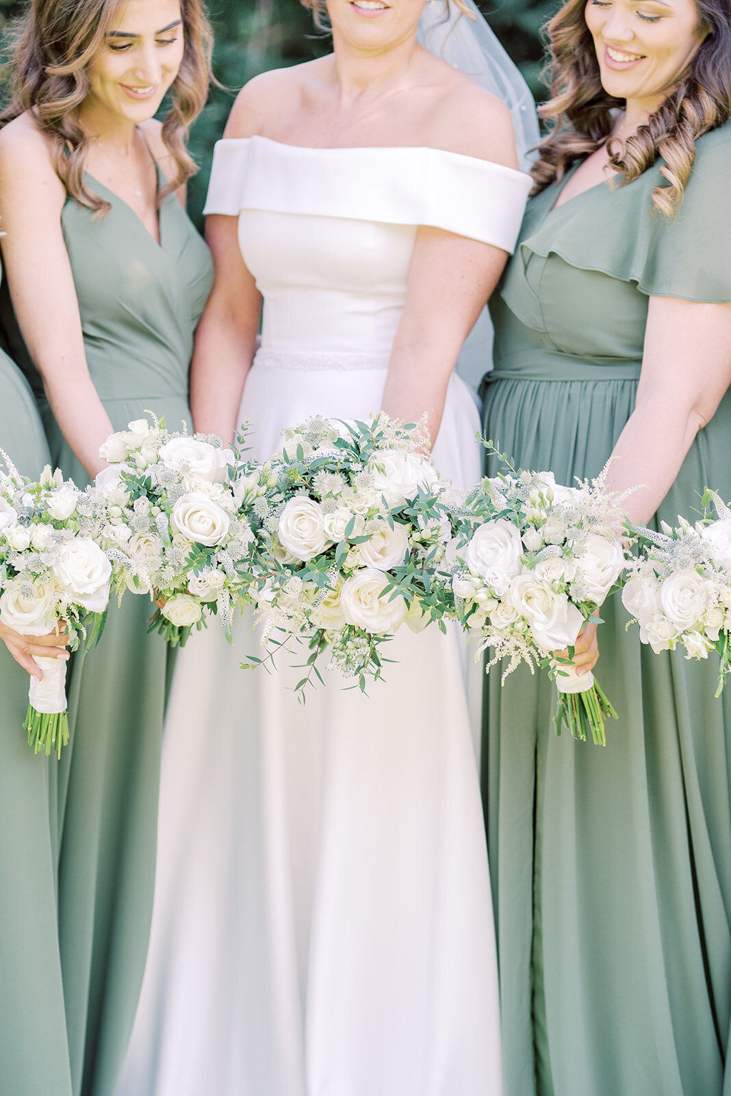 Bride and her bridesmaids, the bride is wearing a white satin dress and the bridesmaids are in sage green dresses, they are holding their bouqets out in front of them and image focuses on the bouquets. the flowers are white roses with eucalyptus