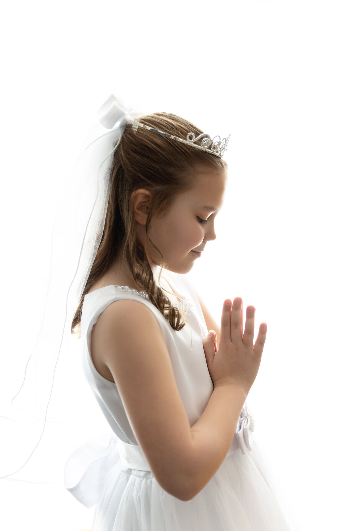 A young girl in a white dress and tiara prays in a white roomNew Jersey Communion Portrait Photographer