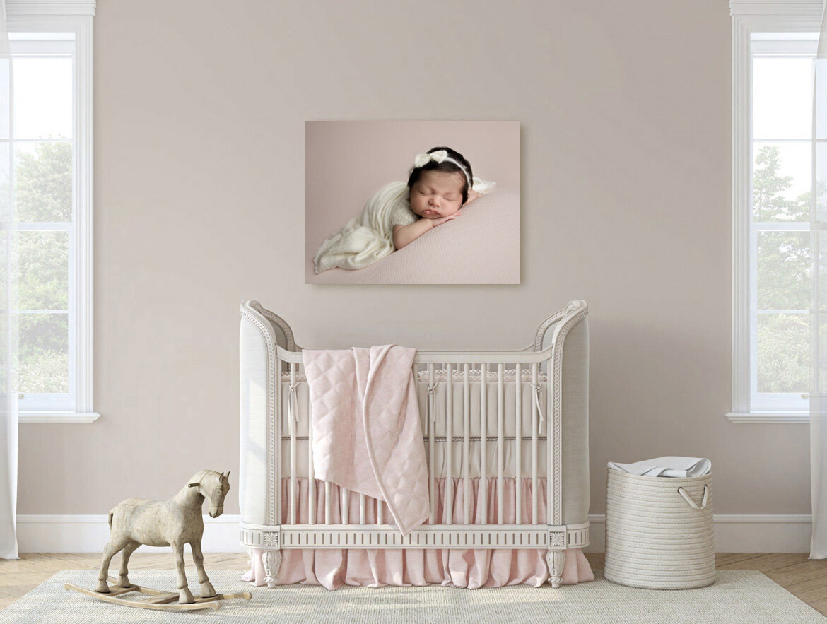 Beautiful pink and white nursery decorated with a gorgeous canvas art piece aover the crib of a newborn baby posed on her tummy faceing towards the camera. She is wearing a simple white bow and a  soft knit romper