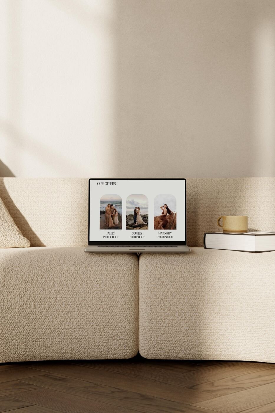 Open laptop on cream boucle sofa showing a photographer’s website offers for family, couples and maternity photoshoots, with coffee cup and book beside it.