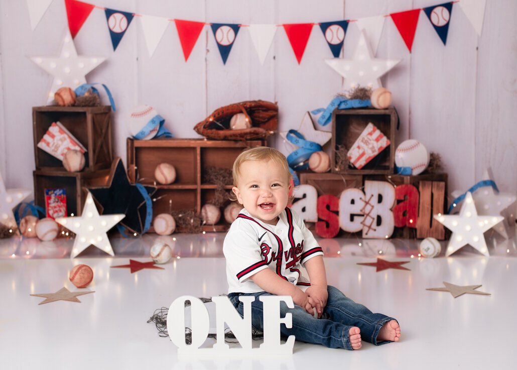 Cake Smash photography -- Smiling one-year-old boy in a Braves jersey sits in a baseball-themed studio set with bats, balls, and red, white, and blue decorations