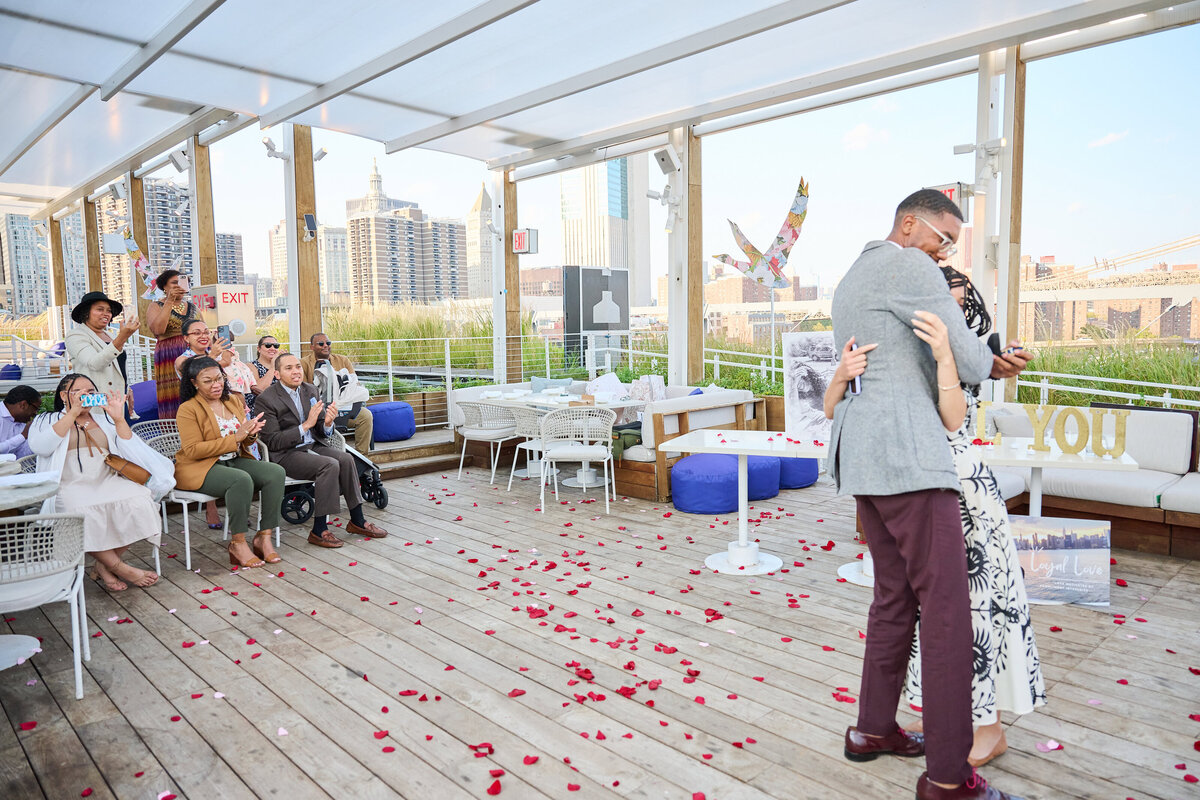 Couple embrace on the Rooftop of the Greens after successful Proposal with friends and family in view.  Brooklyn bridge can be seen in the background.  Taken by Proposal Photographer Corey Lamar Photography NYC