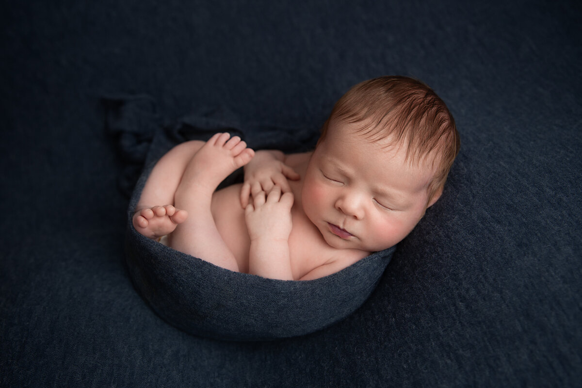 Newborn baby sleeping curled up on his back and wrapped in a navy blue wrap.