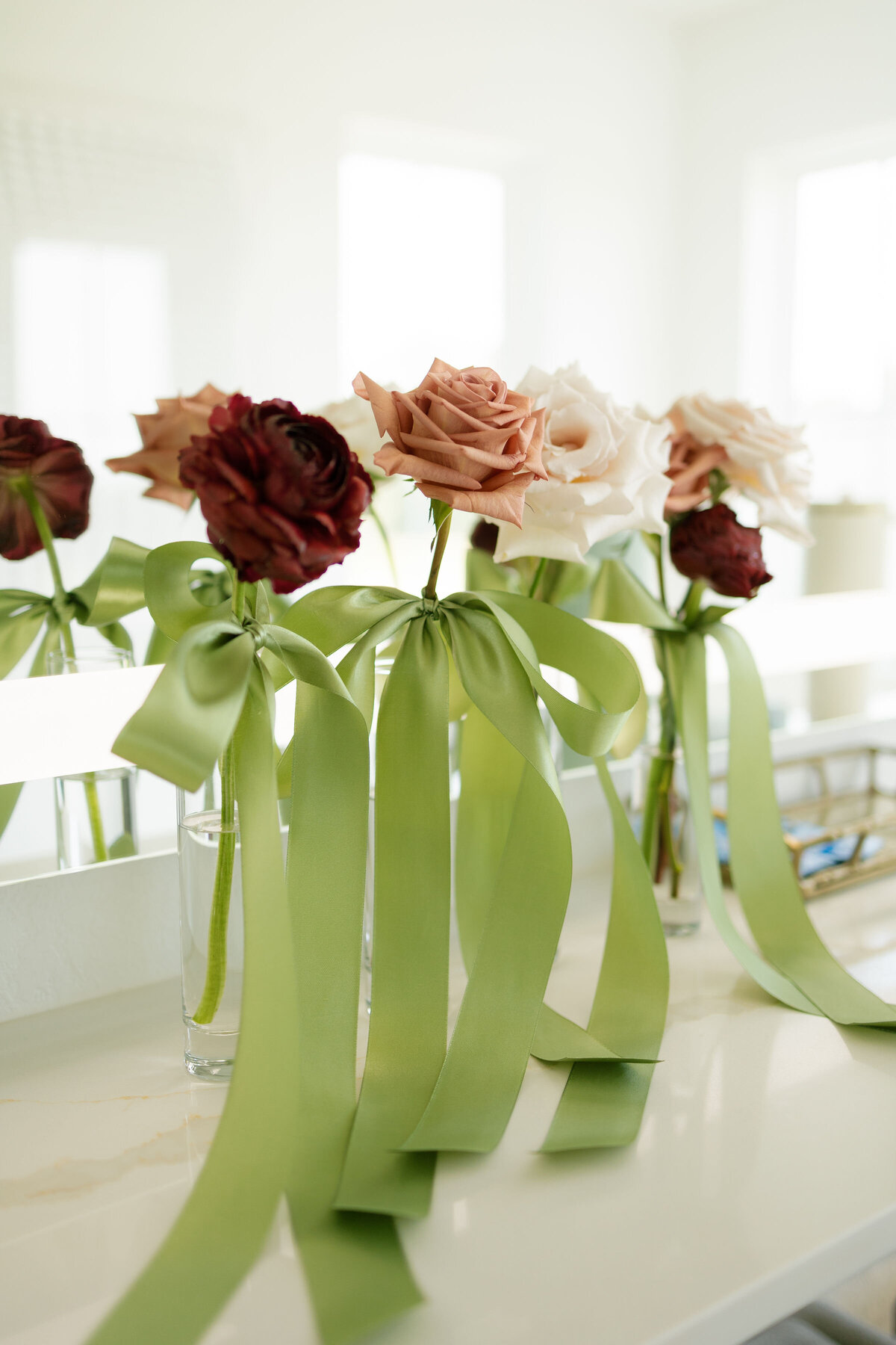 Single long-stem roses in shades of mauve, blush, and burgundy displayed in clear glass vases with flowing green satin ribbons, captured as elegant bridal getting-ready floral styling.