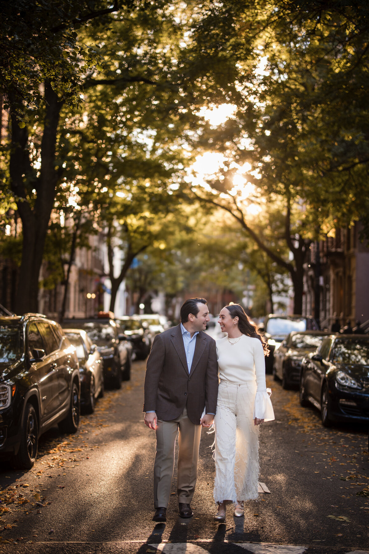 A couple walks hand in hand down a tree-lined street at sunset, smiling at each other at their Soho engagement session.