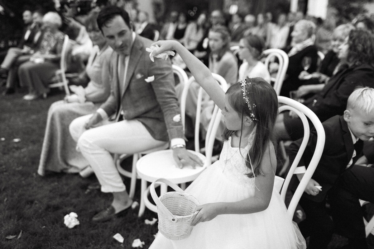 A young girl in a white dress scatters flower petals from a basket at an outdoor event, captured beautifully by an Muskoka wedding photographer. People are seated in the background, adding to the serene charm of the moment.