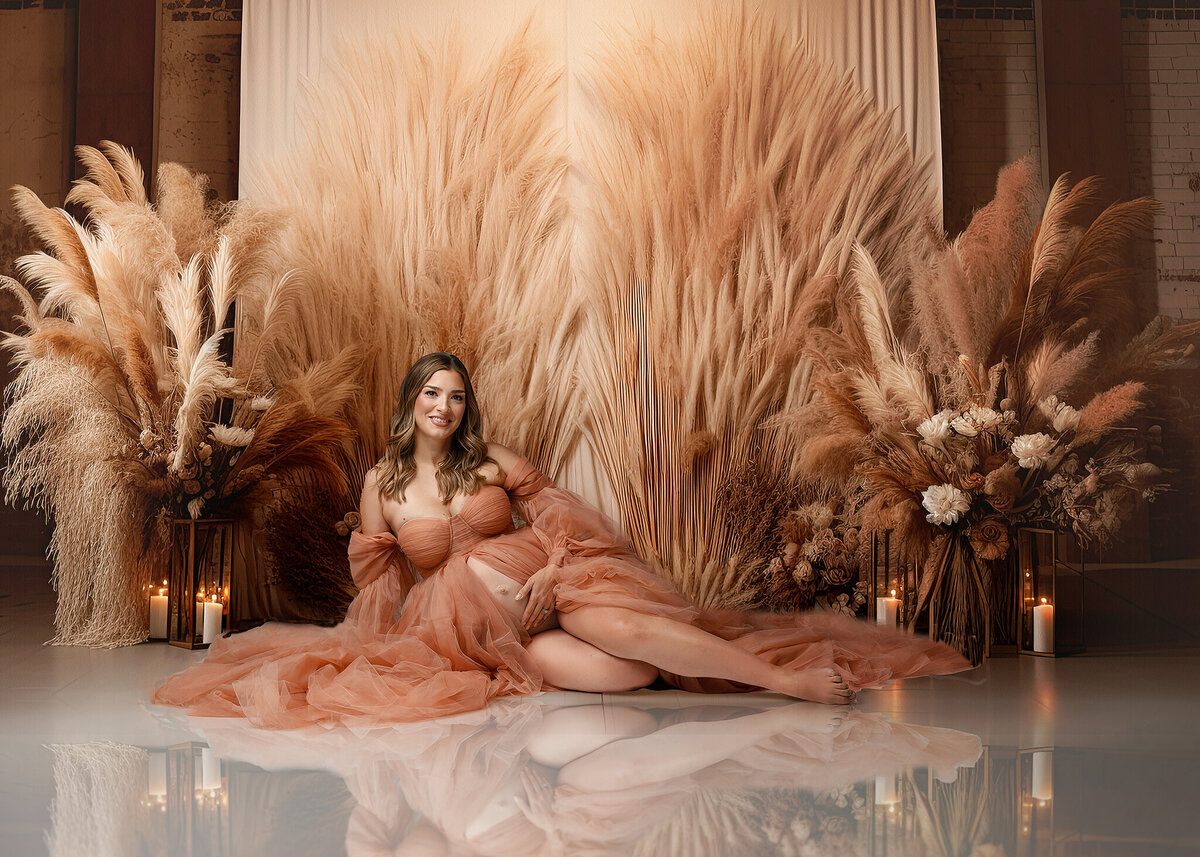 smiling pregnant woman sitting down in a vintage pink gown, studio background wiht pampas grass and textures wiht mirror reflection on the floor