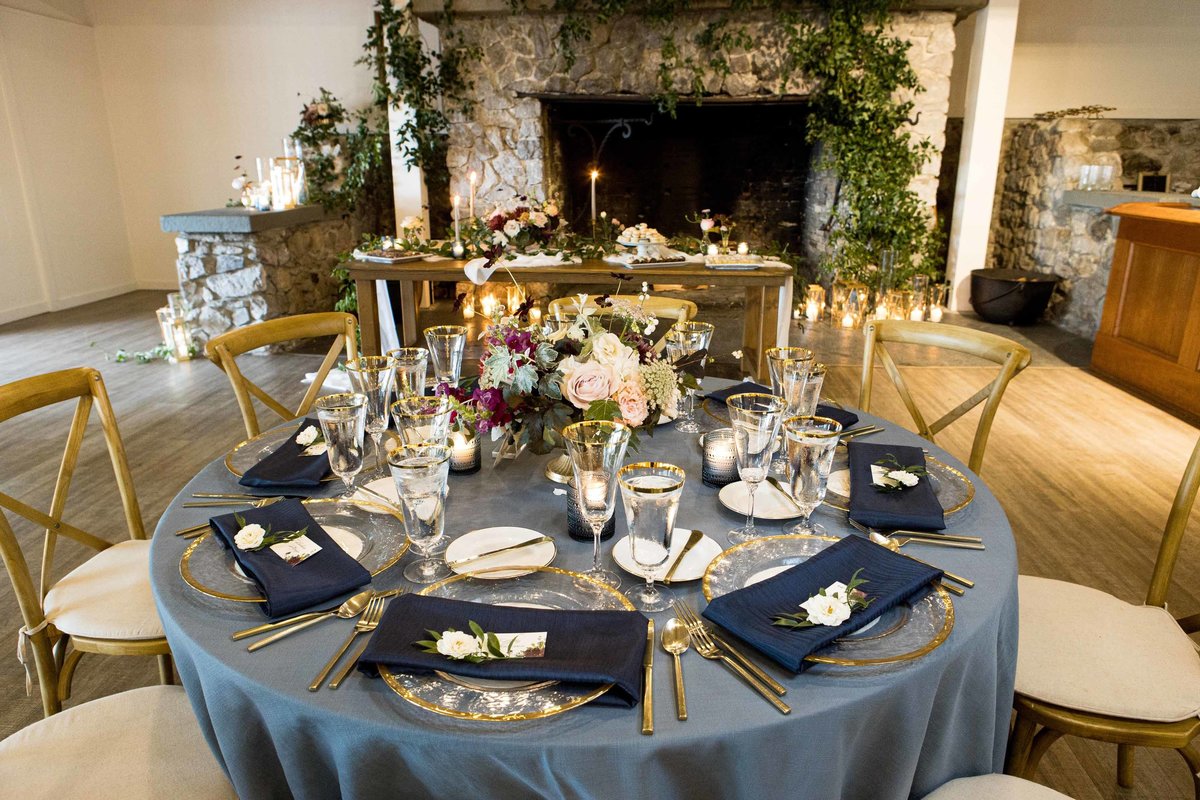 round wedding table in French blue linen, navy napkins, in front of large stone fireplace covered in greenery