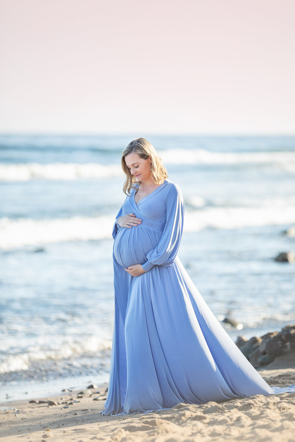 Mom to be in a beautiful pastel blue dress photographed looking at her baby bump in Malibu by the blue ocean