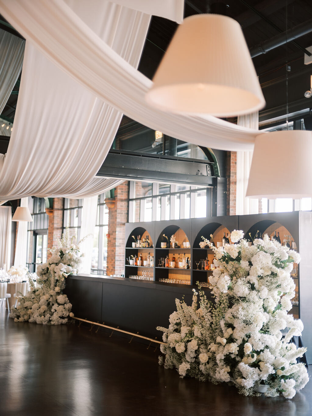 A sleek black bar framed by lush white floral arrangements and draped white fabric at the reception.