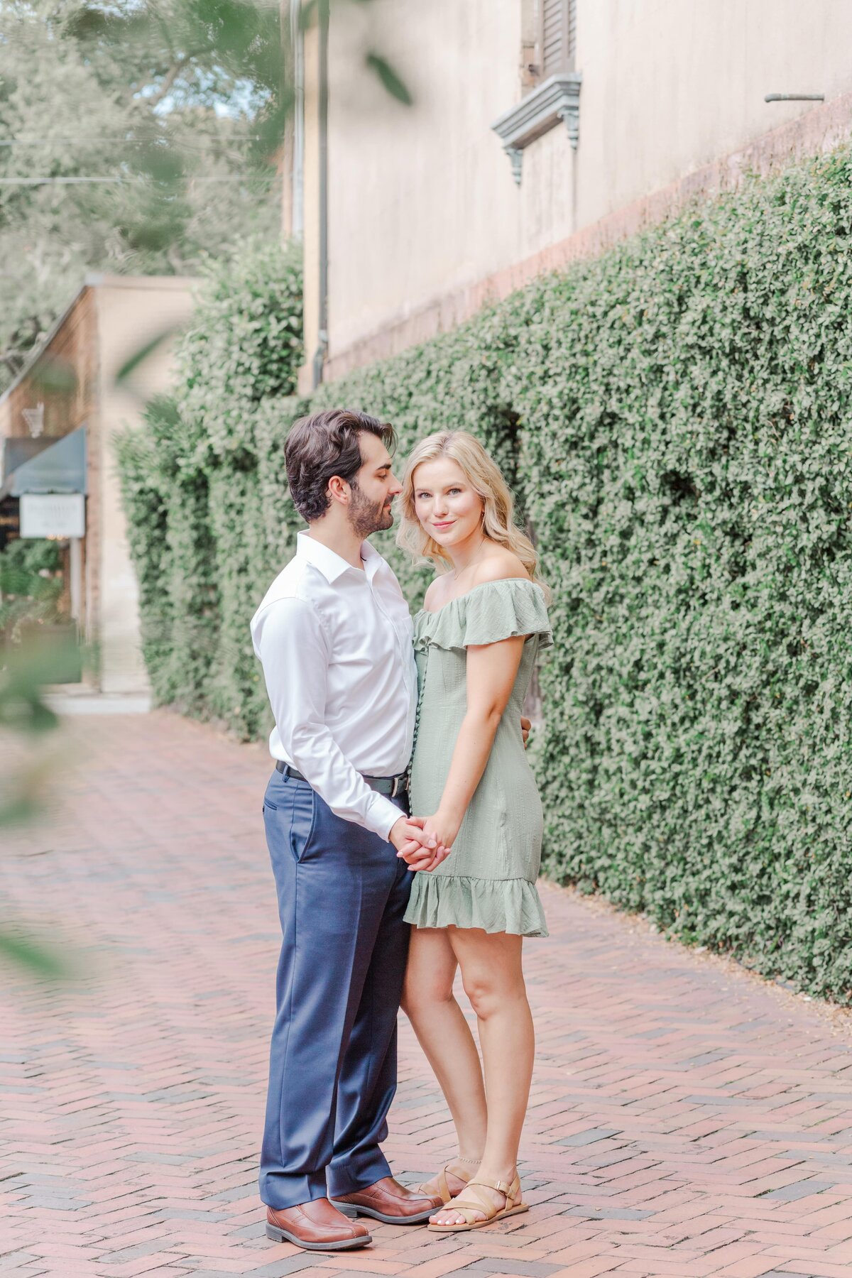 An engaged couple hold hands and embrace on brick sidewalk.