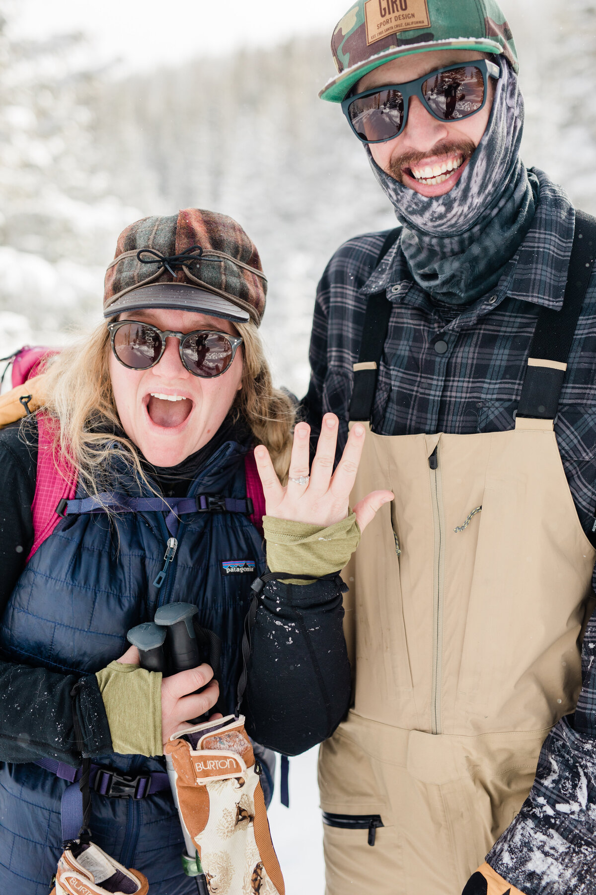 A woman in all backcountry gear excitedly shows off her new ring with her fiance smiling behind her