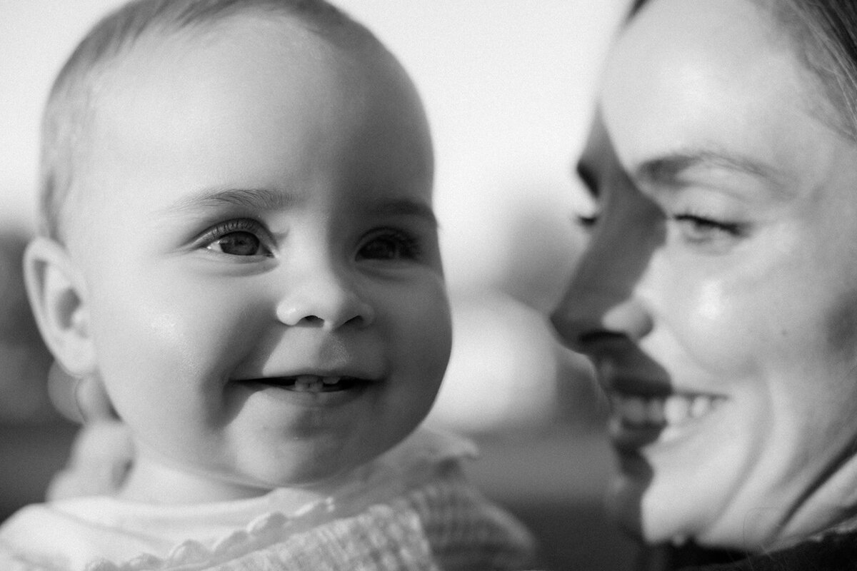 Infant smiles while soaked in sunlight as mother smiles at infant.