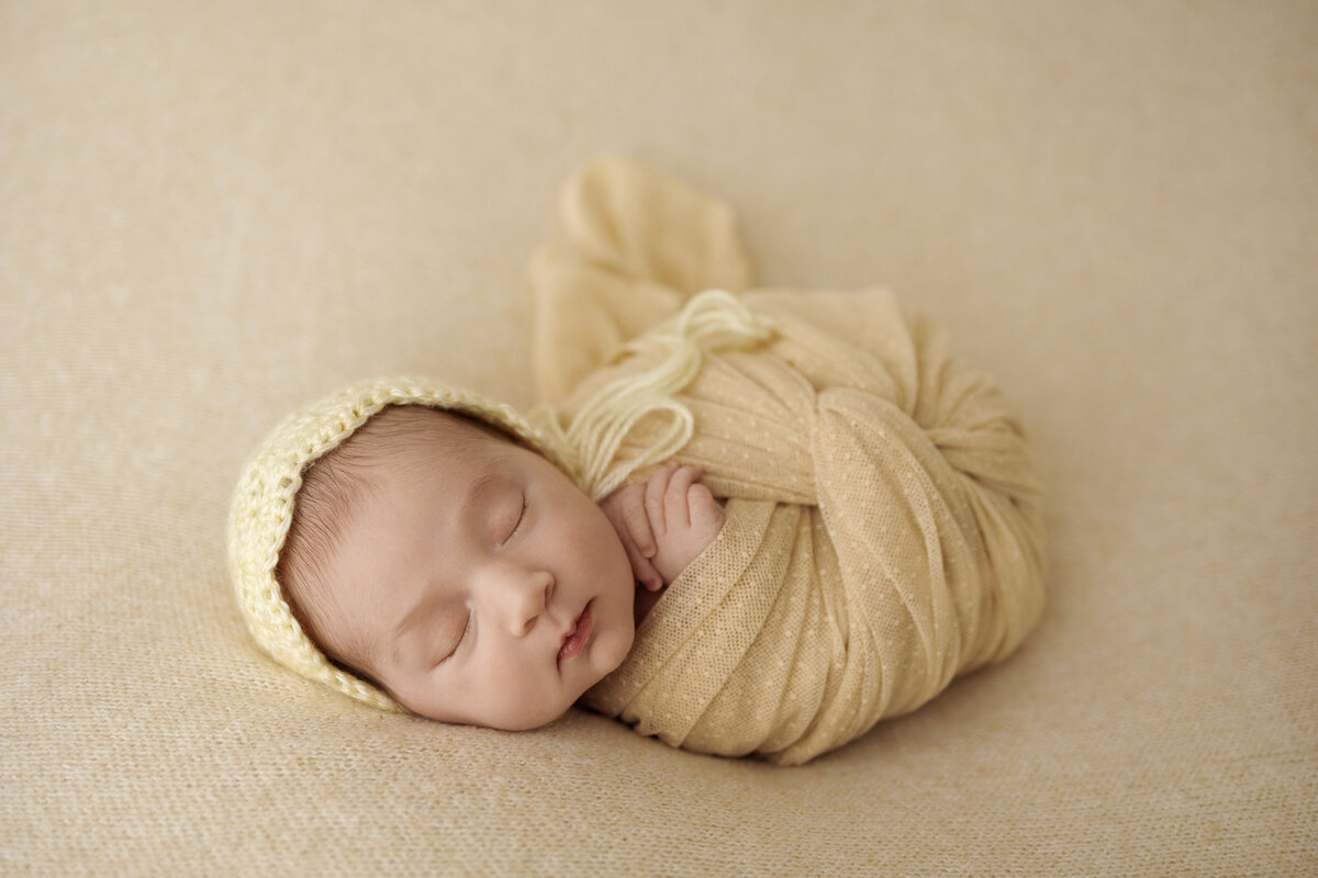 Newborn baby wrapped in a yellow blanket and wearing a matching knit bonnet, peacefully sleeping on a beige background.