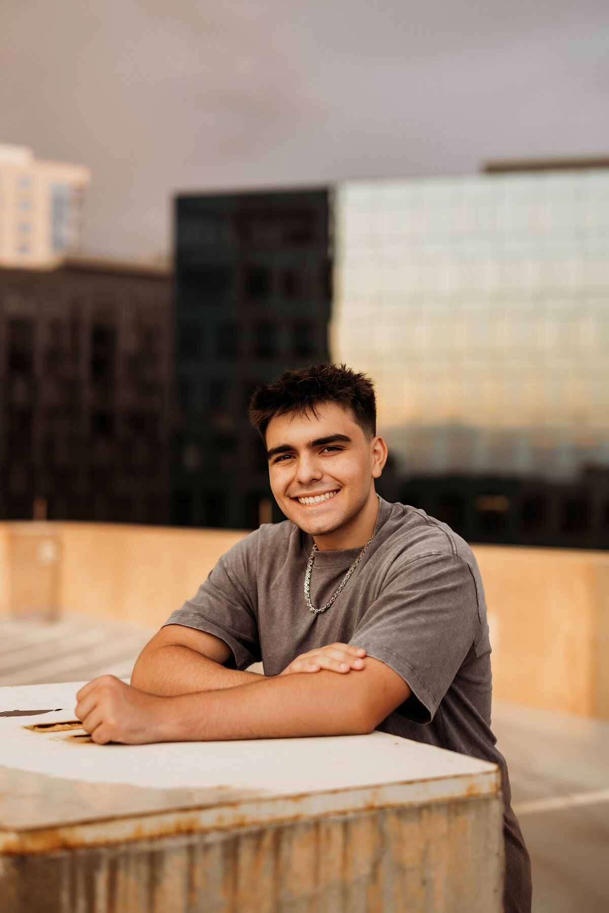 Senior boy leans over concrete pillar with large buildings behind him for his senior photos session