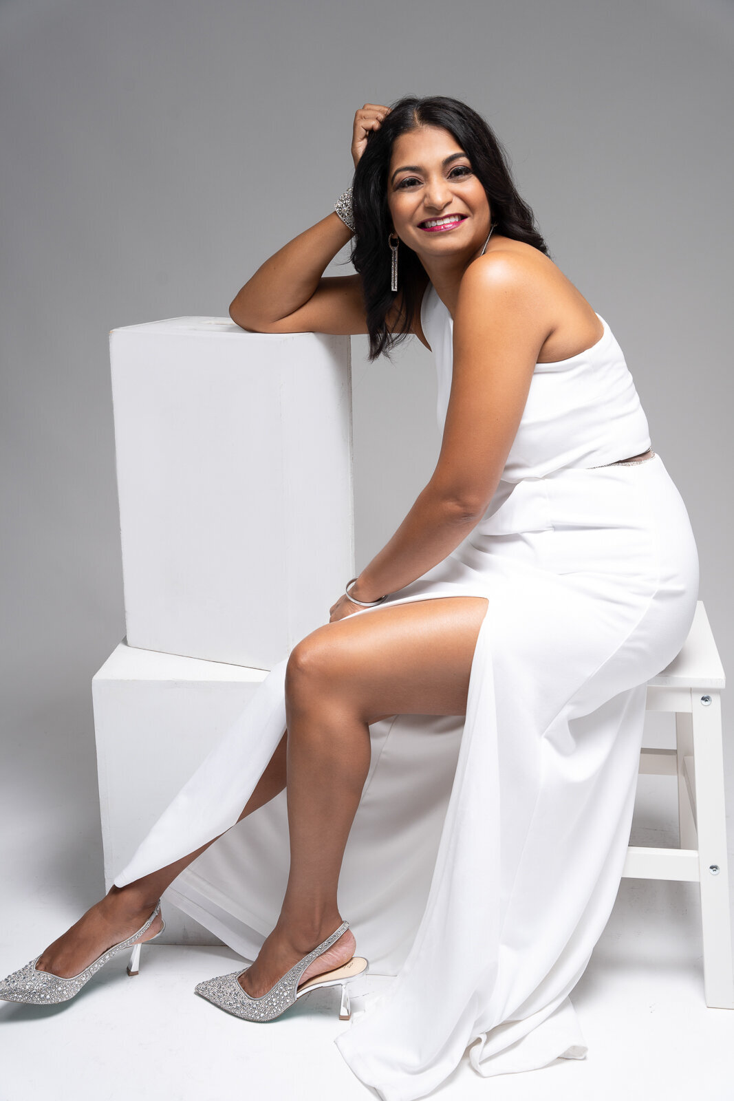 A picture of a woman smiling with a white dress with her arm on white boxes and siting on a white chair