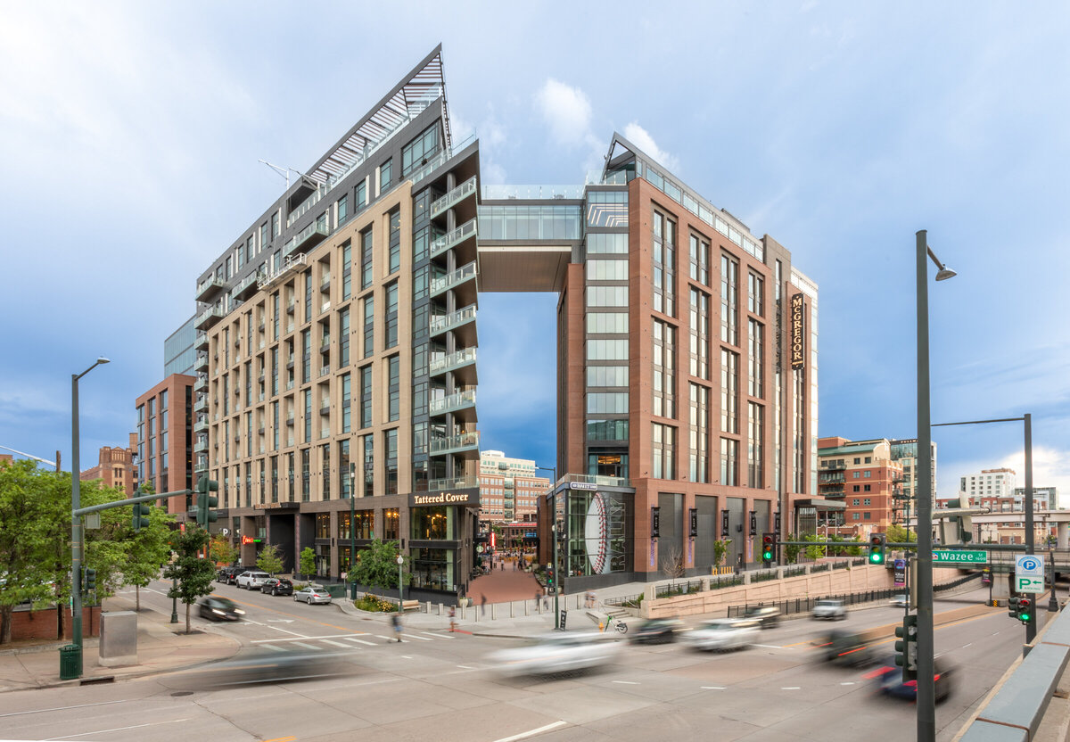 An exterior photo showing the entirety of McGregor Square in Downtown Denver, Colorado. Traffic is rushing past in front and people can be seen walking on the sidewalk.