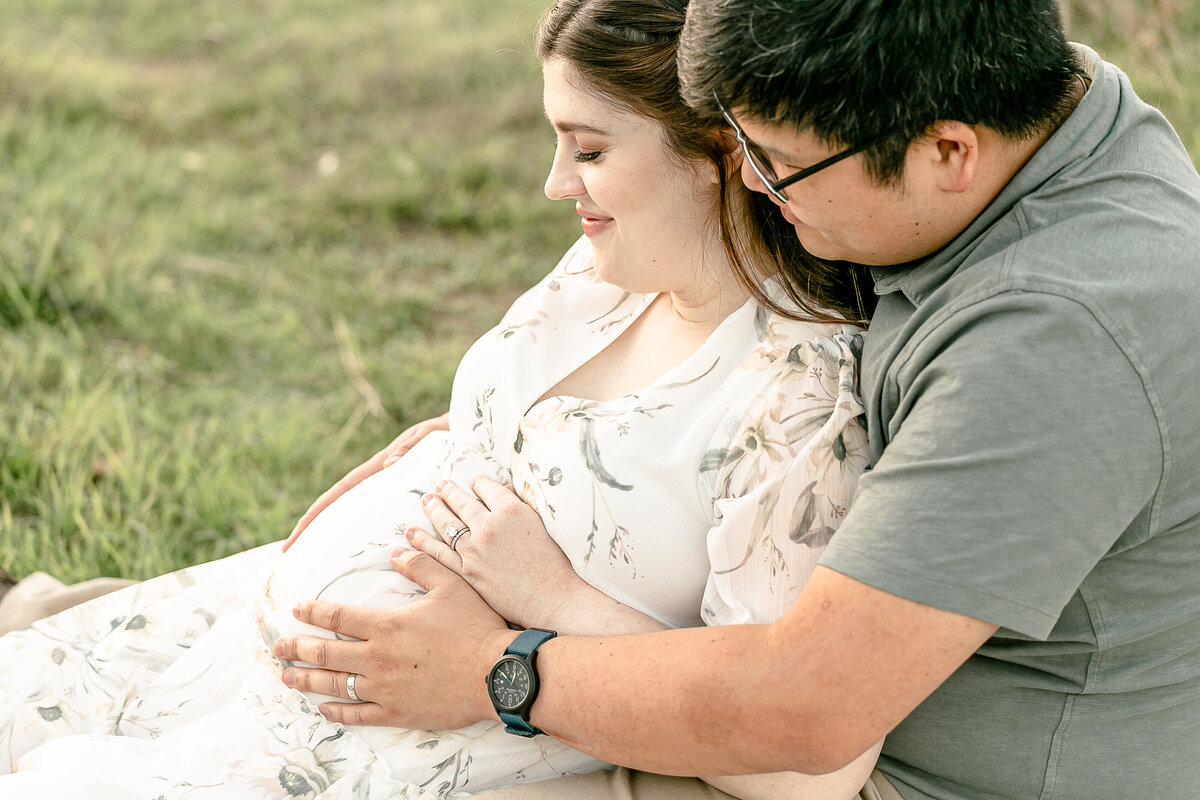 pregnant woman sitting down with man sitting behind her both looking at her belly and resting their hands on her belly. Couple wearing light neutral colors at their outdoor photo session with a light and airy Portland Maternity Photographer.