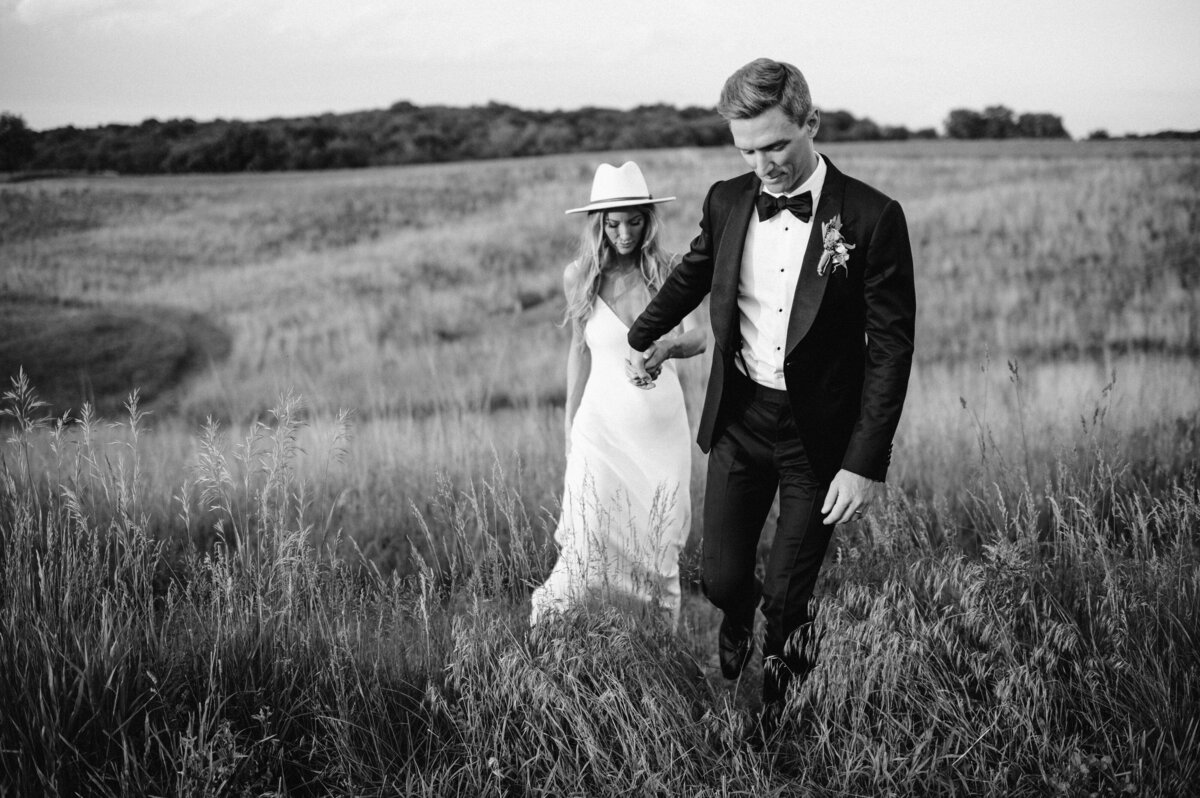 Black tie groom and bride wearing a grace loves lace gown and a hat walk hand and hand through a field in black and white. Photo by Anna Brace, a des moines wedding photographer.