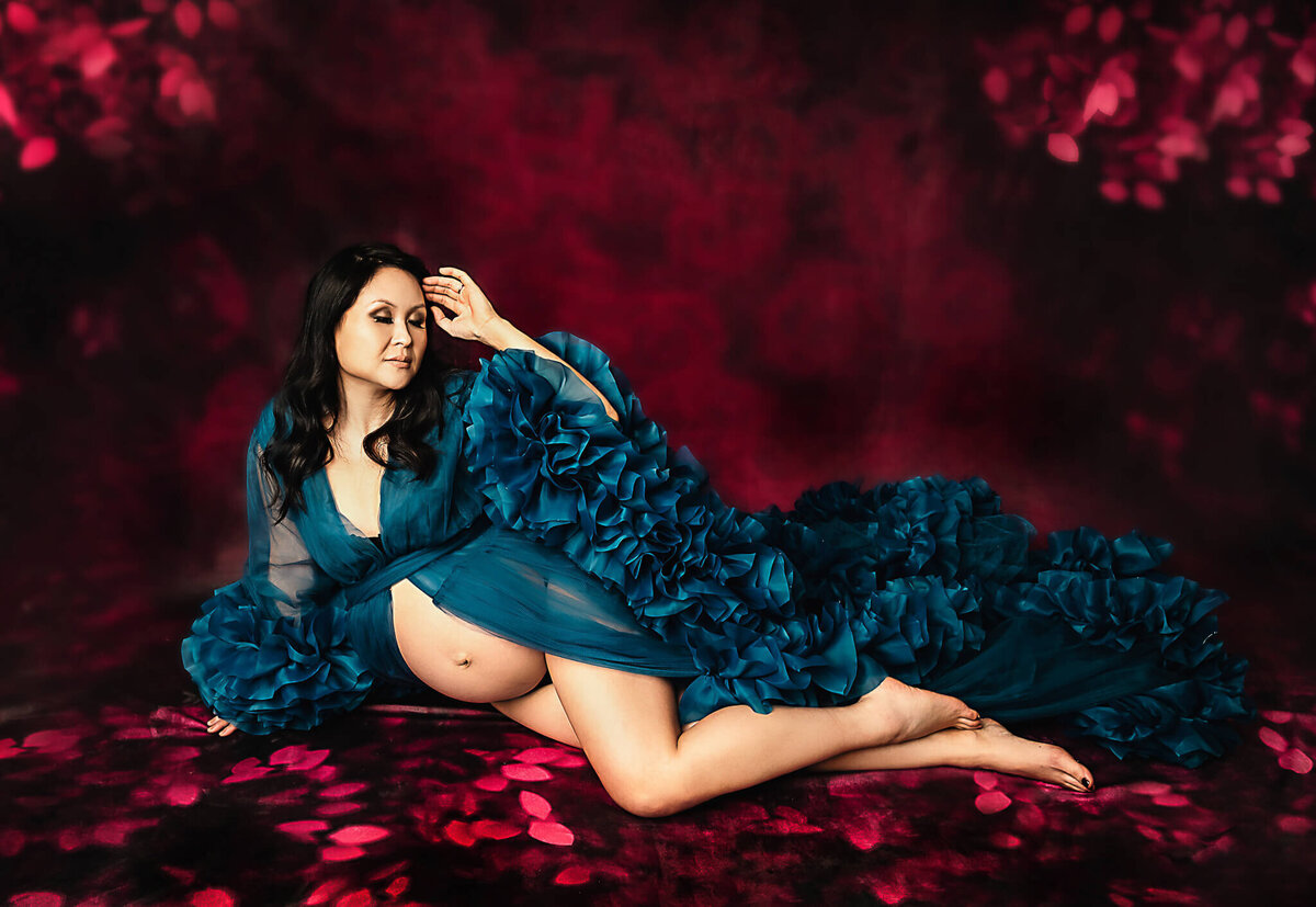 Stunning pregnant mom laying on her side on a red floral backdrop with bare belly exposed wearing a ruffled royal blue robe.