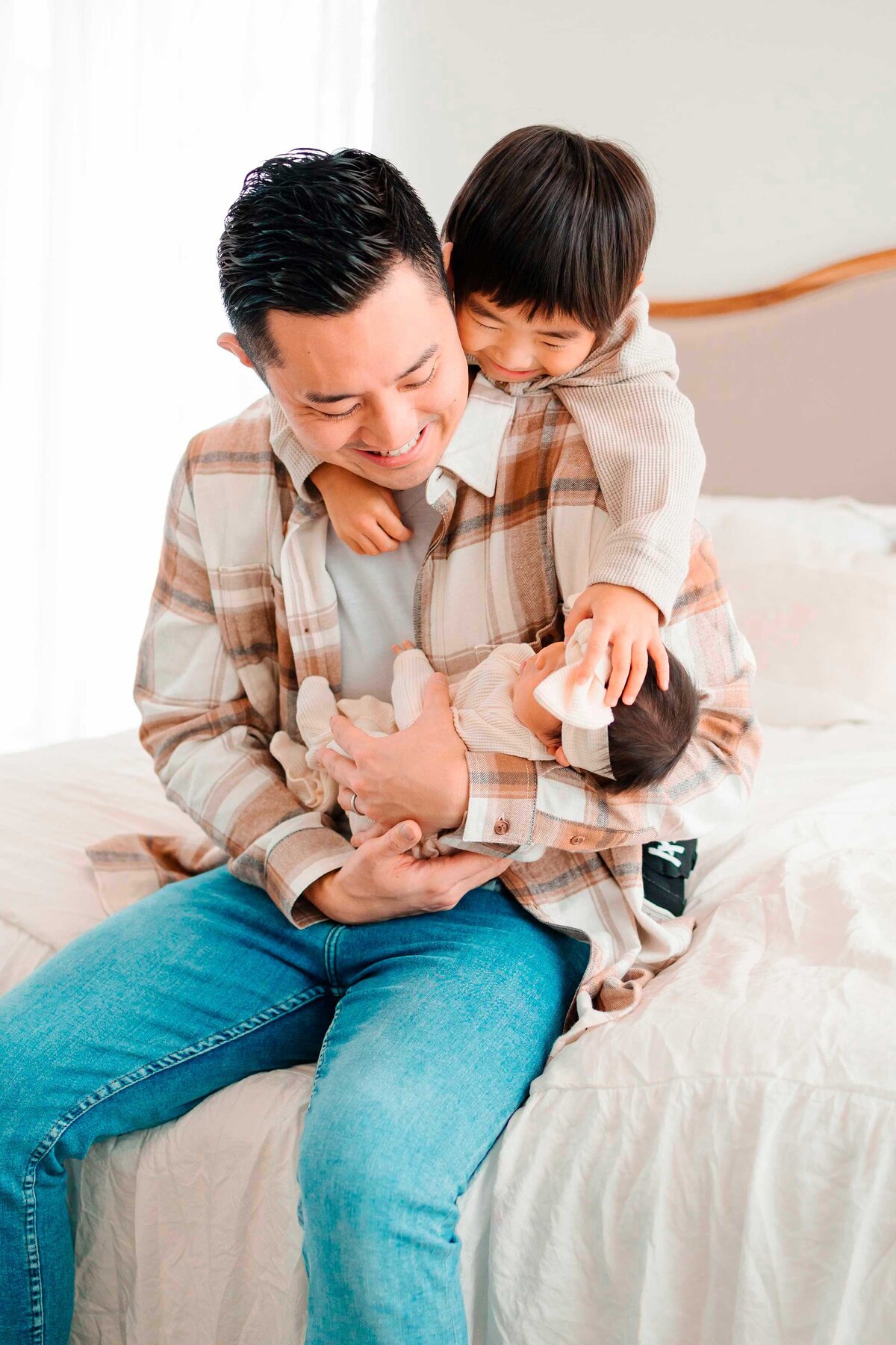 A joyful father sits on a white bed with his two little boys, all expressing happiness with the newborn baby girl.