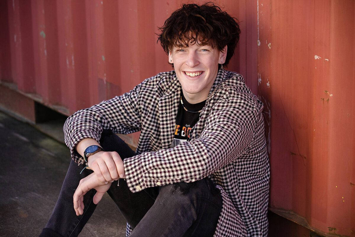 Seated senior portrait in front of a red cargo container at Camp North End, styled in a Beatles tee and checked shirt by Charlotte senior photography studio.